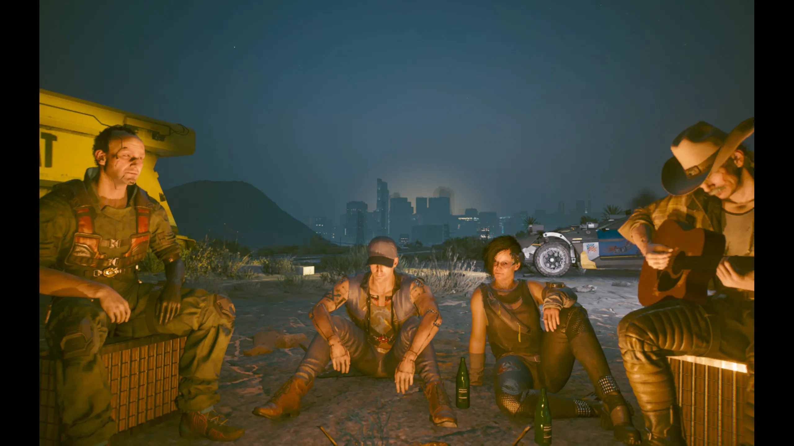 Four members of the Aldecaldos nomad clan relax around a campfire in the desert at dusk, lit in warm orange against a deep blue sky. On the far right a man in a cowboy hat plays an acoustic guitar, two others sit cross-legged in the center with beer bottles in the sand, and a rugged man in armored gear leans against a yellow container on the left. Behind them the glowing skyline of Night City rises in the distance, with a parked vehicle off to the side.
