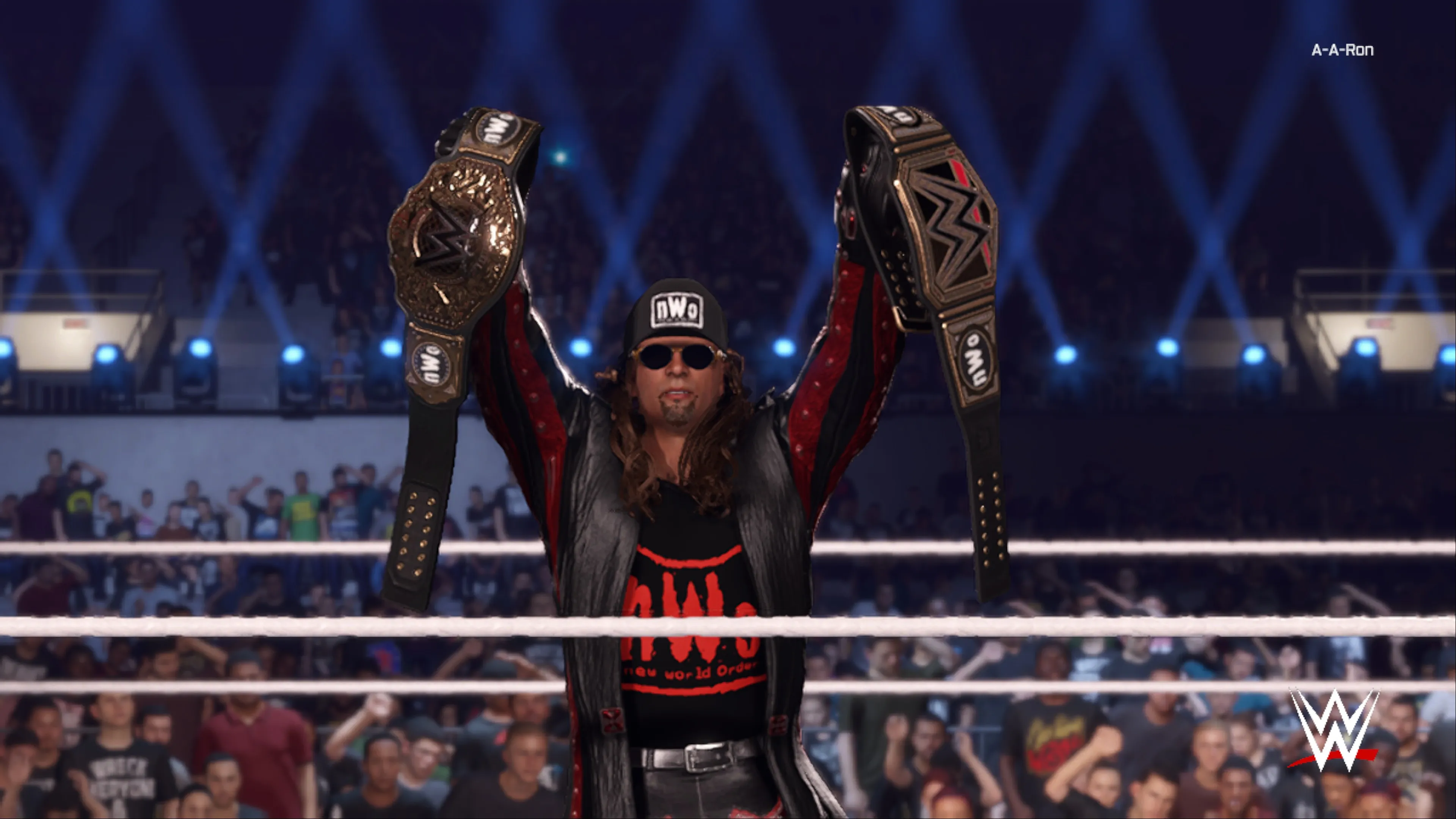 A wrestler in nWo attire and sunglasses celebrates in the ring with two championship belts raised overhead, standing behind the ropes with a crowd and blue stage lighting in the background in WWE 2K25