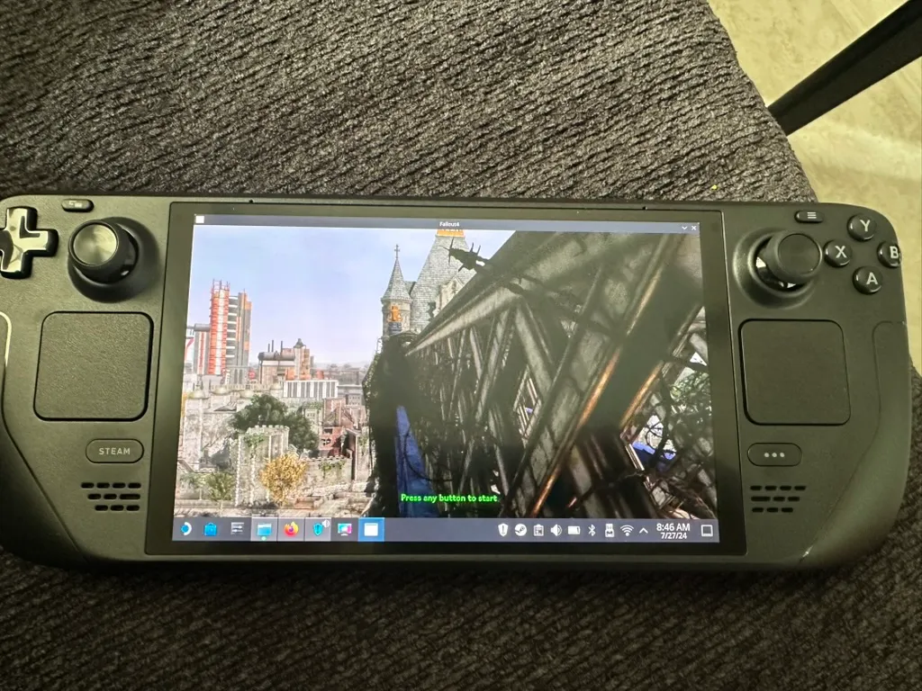 A handheld gaming device, the Steam Deck, is shown on a gray fabric surface. The screen displays the video game, Fallout London, with a scene featuring a detailed, post-apocalyptic cityscape with buildings and a large bridge. The device’s controls, including a directional pad, buttons, and joysticks, are visible on either side of the screen. The device’s interface at the bottom of the screen shows various icons and the time “8:46 AM” with the date “7/27/24.