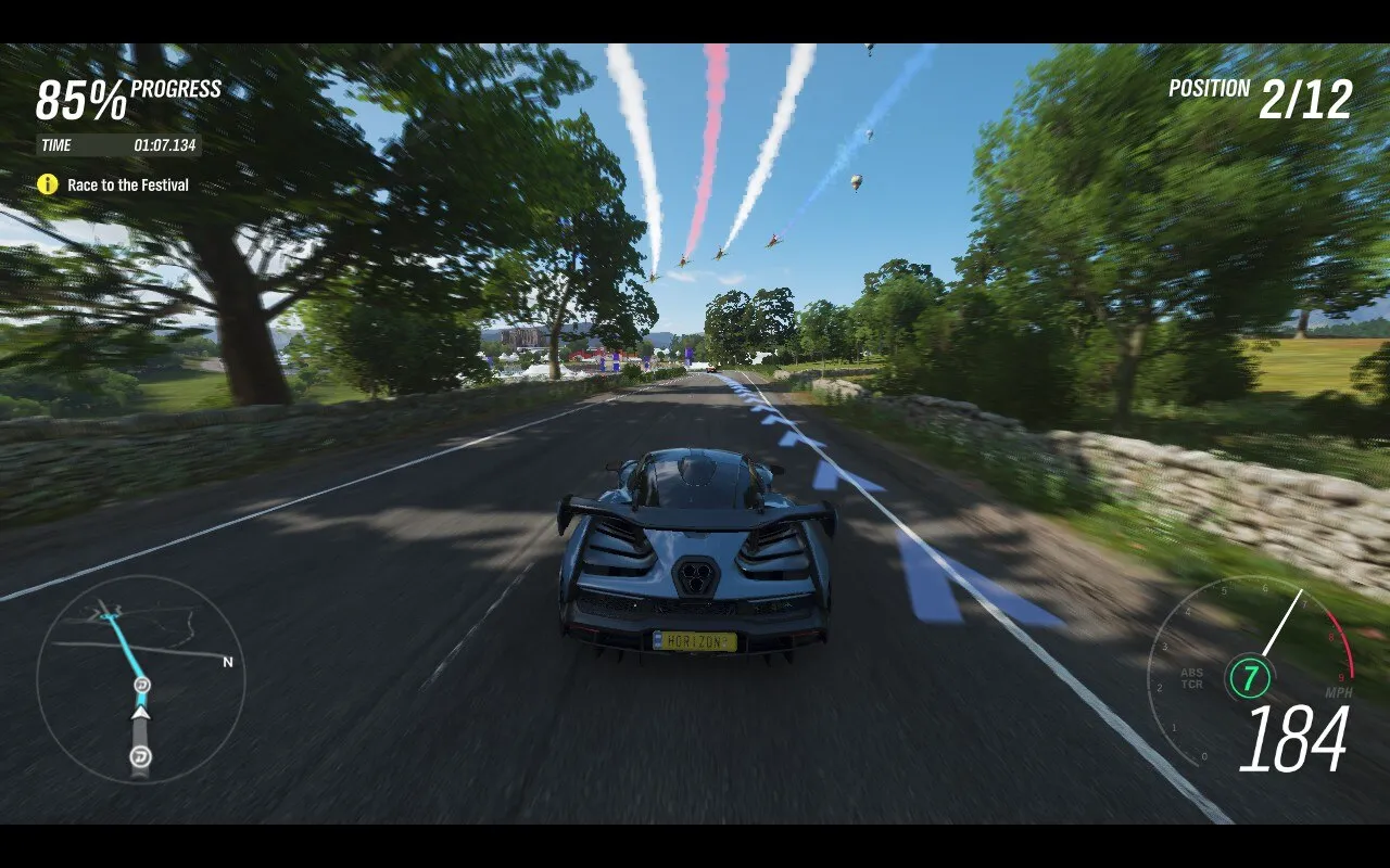 A screenshot from Forza Horizon 4, a racing video game showing a high-speed car race on a scenic, tree-lined road. The player’s car, a sleek black sports car with a license plate that reads “HORIZON,” is in the foreground. The game interface indicates 85% progress in the race, with a time of 1:07.134, and the player’s position is 2nd out of 12. In the sky above, colorful smoke trails from flying aircraft add to the festive atmosphere. A mini-map and speedometer are displayed on the screen, showing the car’s speed at 184 mph.