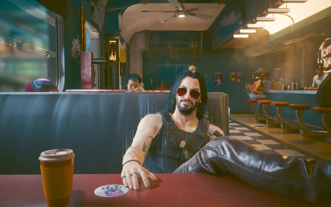 Johnny Silverhand lounges in a red vinyl diner booth, one arm stretched along the seat back and the other resting on the table beside a takeout coffee cup. He wears round tinted sunglasses, a black tactical vest over bare tattooed arms, and dog tags, his long dark hair framing a bearded face. Behind him a retro diner glows with warm neon and hanging lights, with a few patrons at the counter and booths.