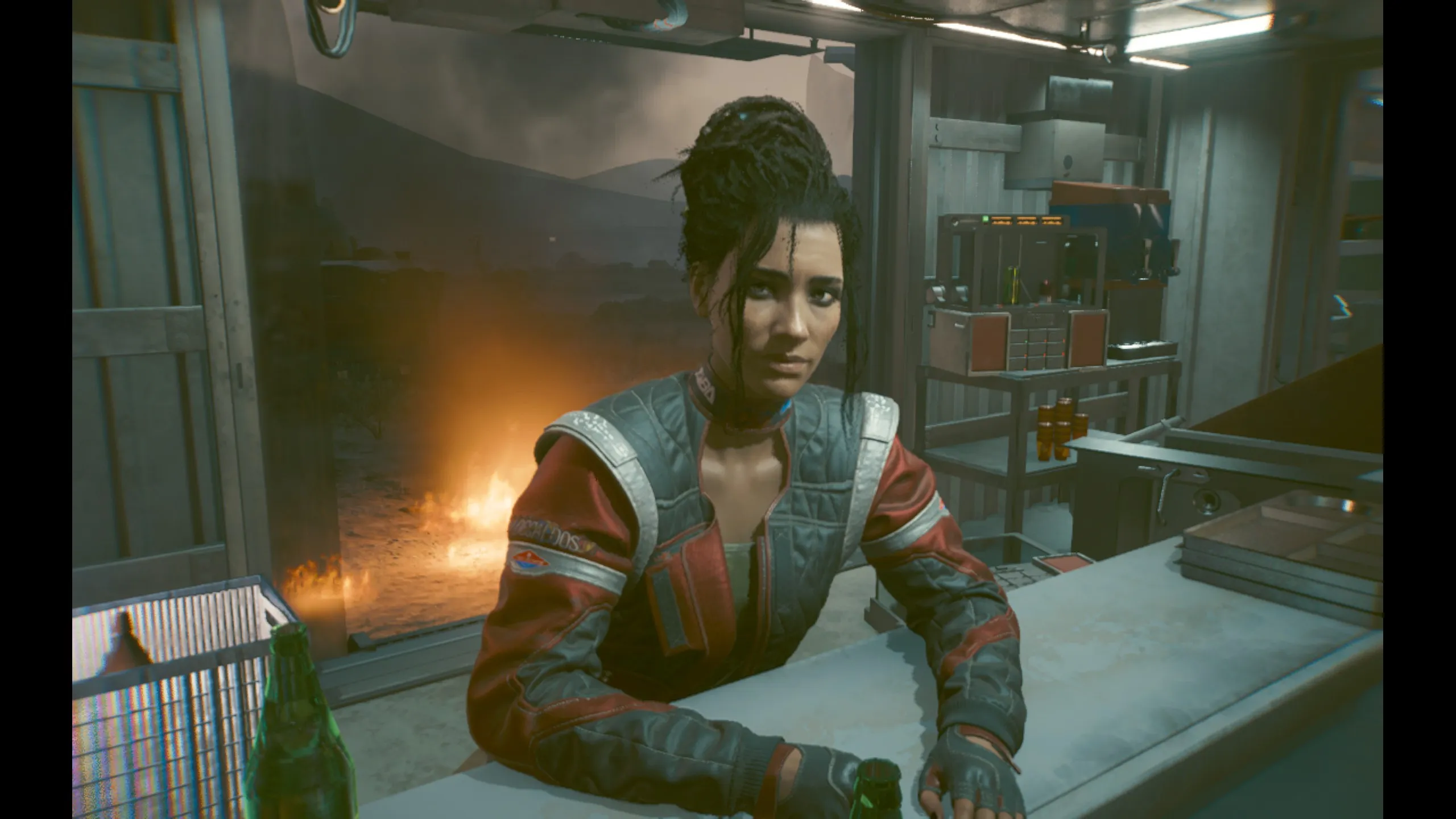Panam Palmer leans on a counter inside a dimly lit nomad trailer, looking toward the camera with a tired, wary expression. Her dark hair is pulled up in a messy topknot with loose strands framing her face, and she wears a red and grey leather Aldecaldos jacket over fingerless gloves. Empty green bottles sit on the counter in front of her, and through a window behind her a campfire burns against a hazy desert landscape at dusk.