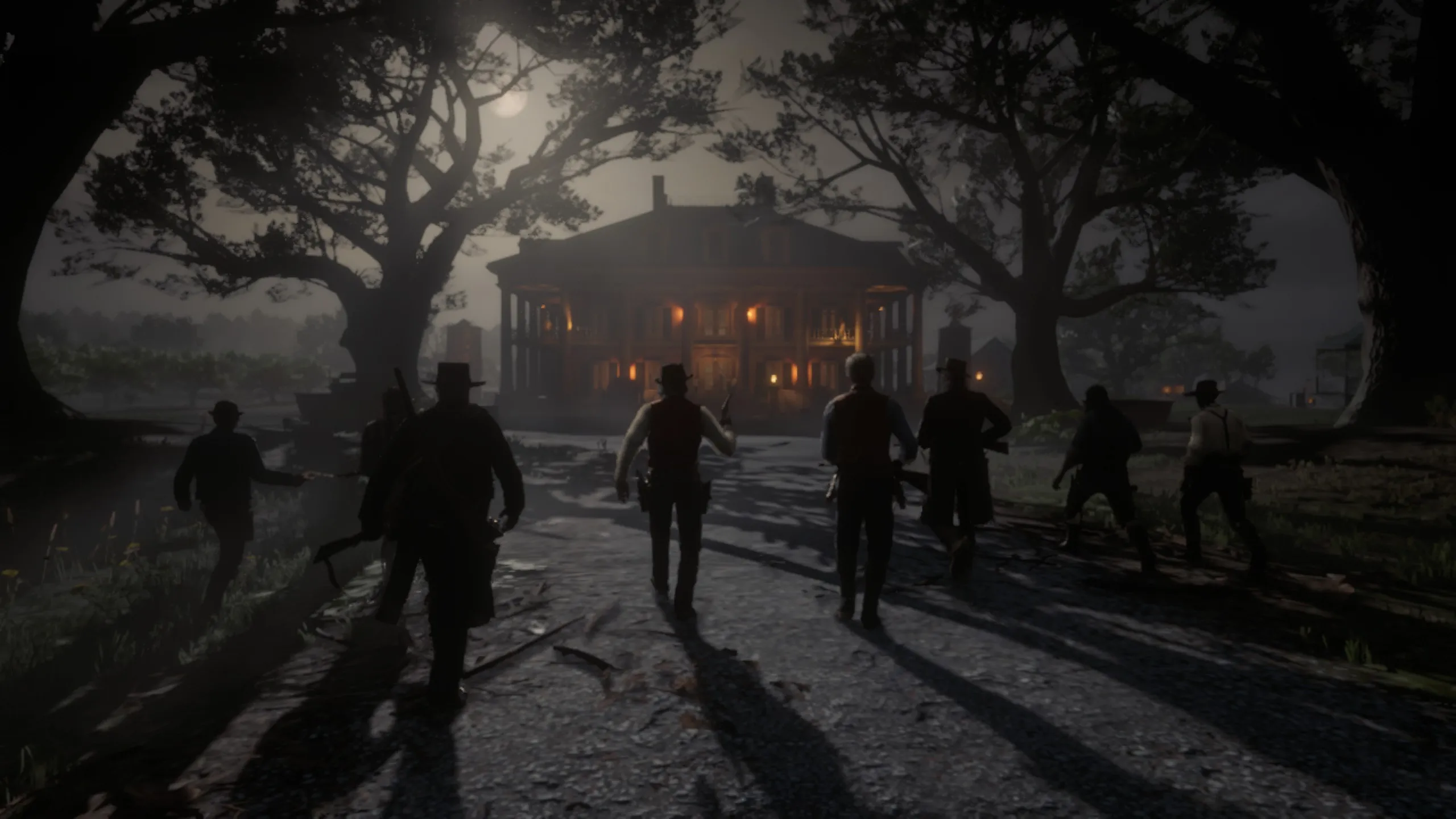A group of armed figures walks towards a large, illuminated mansion at night in a cinematic scene from Red Dead Redemption 2. The moonlight casts long shadows on the ground, while towering trees frame the path leading to the grand house. The mansion’s warm lighting contrasts with the dark surroundings, creating a tense and dramatic atmosphere.