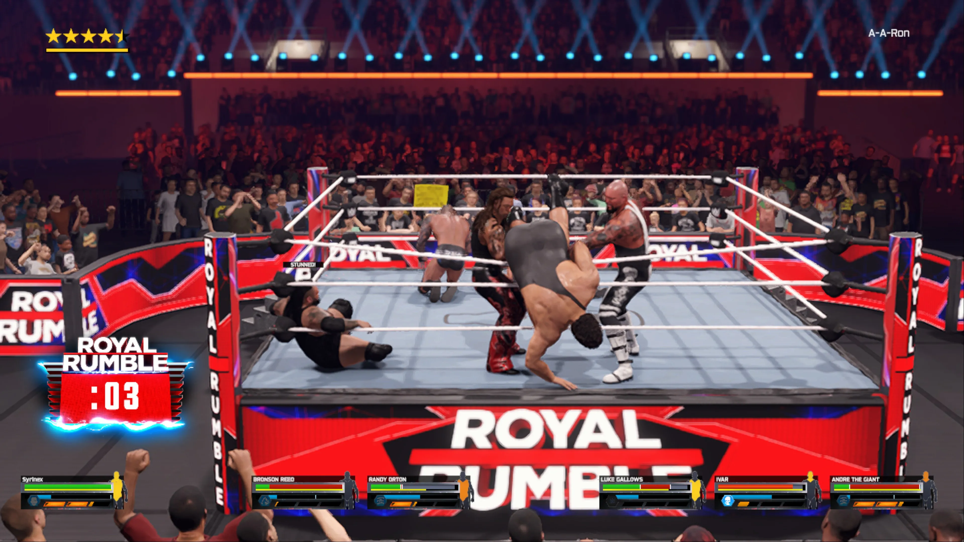 Multiple wrestlers battle in the ring during a Royal Rumble match with one wrestler performing a diving move while others are down on the mat, surrounded by a packed arena crowd and Royal Rumble branded ring apron with a match timer showing 03 seconds until the next entrant in WWE 2K25